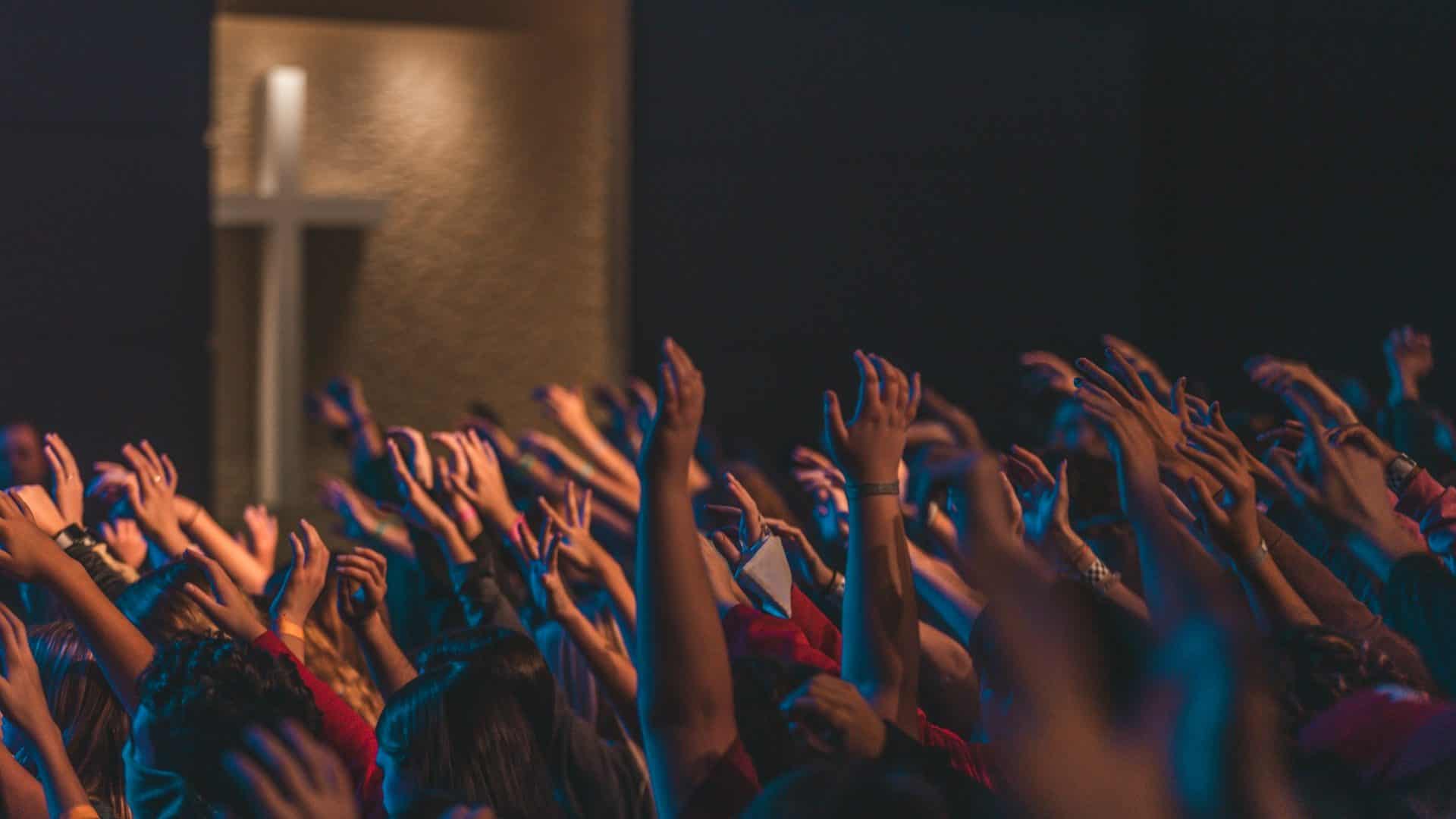 A congregation seen from behind, hands raised before a lit cross