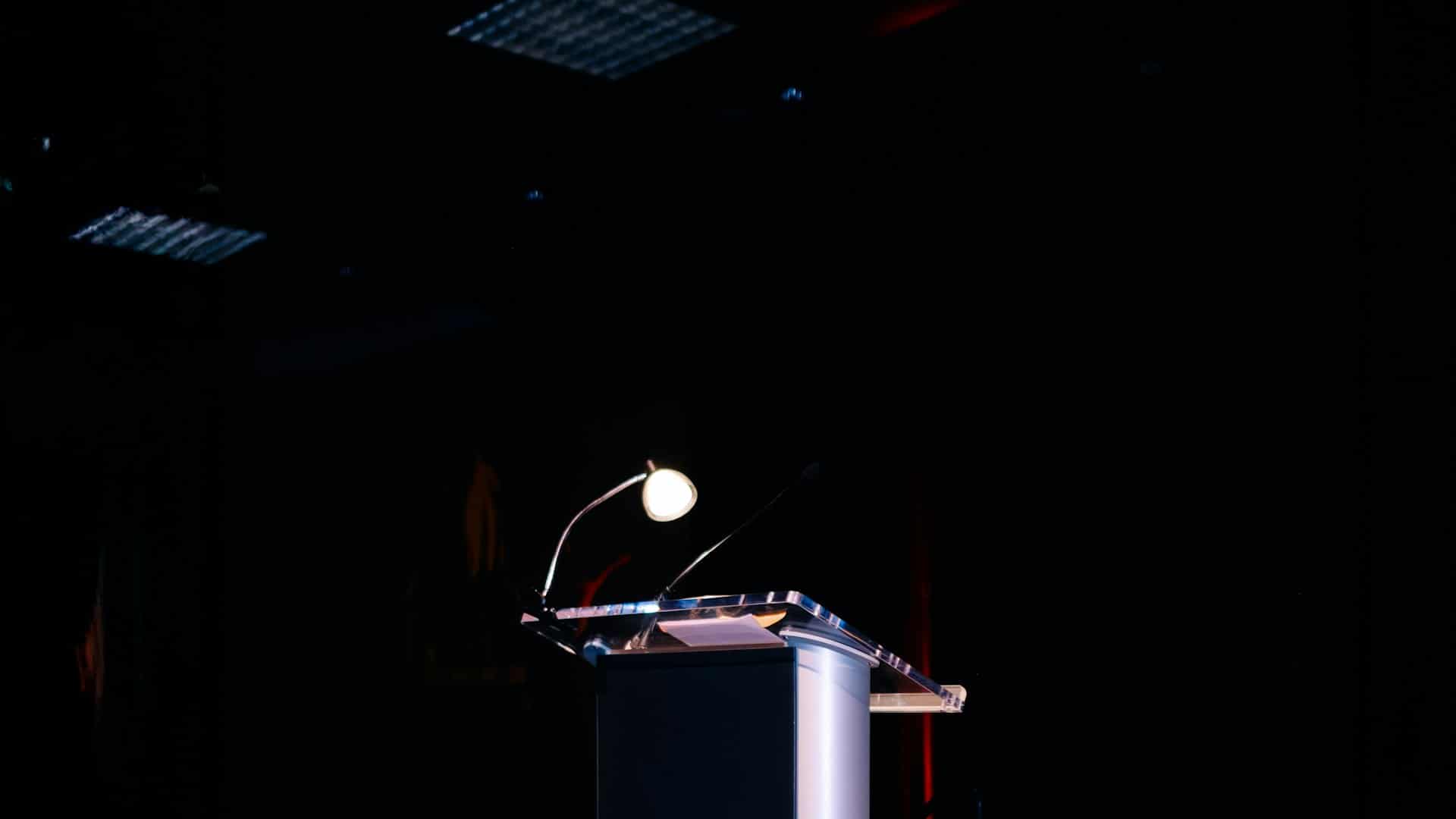 A lectern lit by a lamp, microphone in place, in a dark room